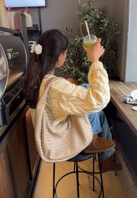 Maglione a maglia crema, borsa beige intrecciata e stivali marroni. Bevanda verde in bicchiere trasparente con cannuccia. Ambiente da caffè interno con piante.