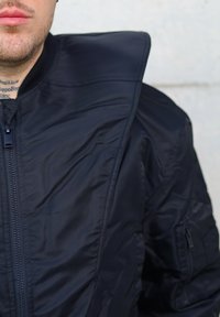 Veste bombardier noire avec un col montant, texture matelassée et fermeture éclair à l'avant avec une poche latérale supplémentaire.