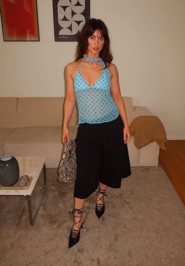 Žena stojaca v interiéri, oblečená v modrom bodkovanom halter top, čierne široké nohavice, čierne šnurovacie lodičky a drží kabelku zo zvieracej kože s hadím vzorom.