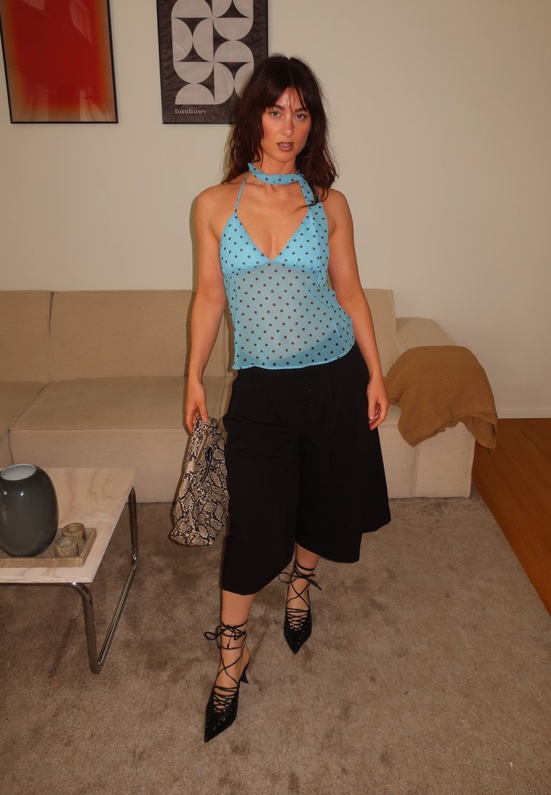 Femme debout à l’intérieur portant un haut halter bleu à pois, un pantalon large noir, des talons lacés noirs, et tenant un sac en peau de serpent.