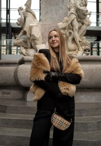 Νεαρή γυναίκα με μαύρο σύνολο, ανοιχτόχρωμη γούνινη εσάρπα και δερμάτινα γάντια, κρ ατώντας μια καρό τσάντα, στέκεται μπροστά σε περίτεχνη πέτρινη βρύση στο εσωτερικό.