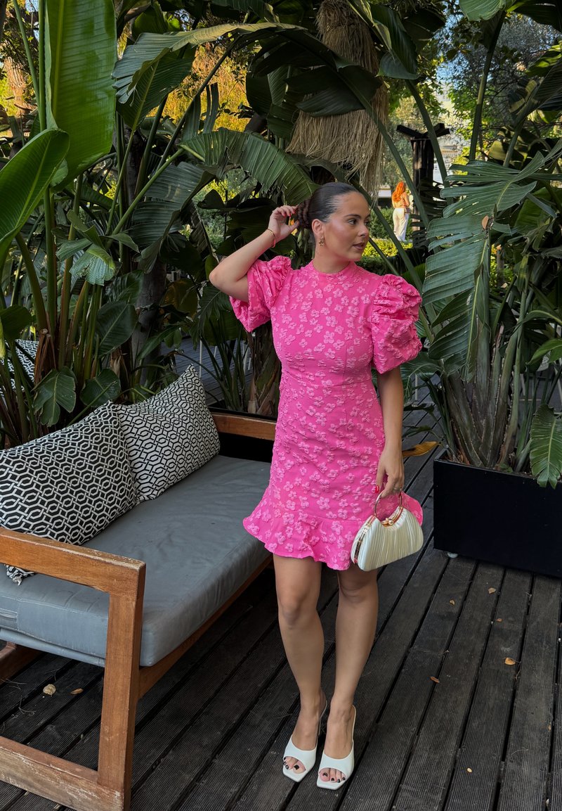 Femme en robe fleurie rose à manches bouffantes et talons blancs se tient sur une terrasse en bois près de plantes tropicales et d’un banc en bois avec des coussins à motifs.