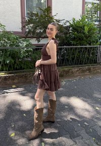 Moteris be rankovių rudos suknelės ir aukštų rudų batų, laikanti šviesiai rudos spalvos rankinę, stovi saulės apšviestame grindinyje šalia žalumos ir pastato.