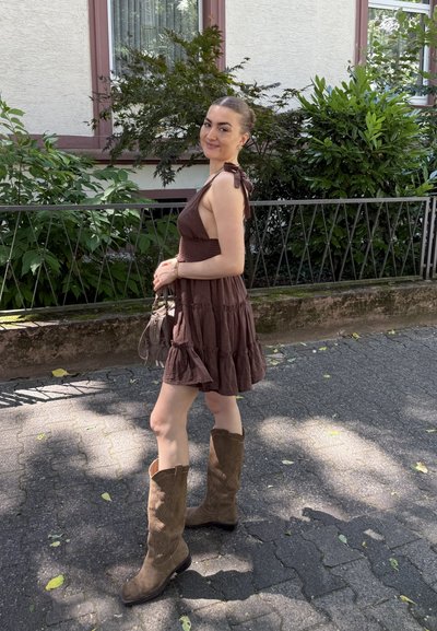 Mujer con un vestido marrón sin mangas y botas marrones altas, sosteniendo un bolso beige, de pie en un camino pavimentado iluminado por el sol cerca de vegetación y un edificio.