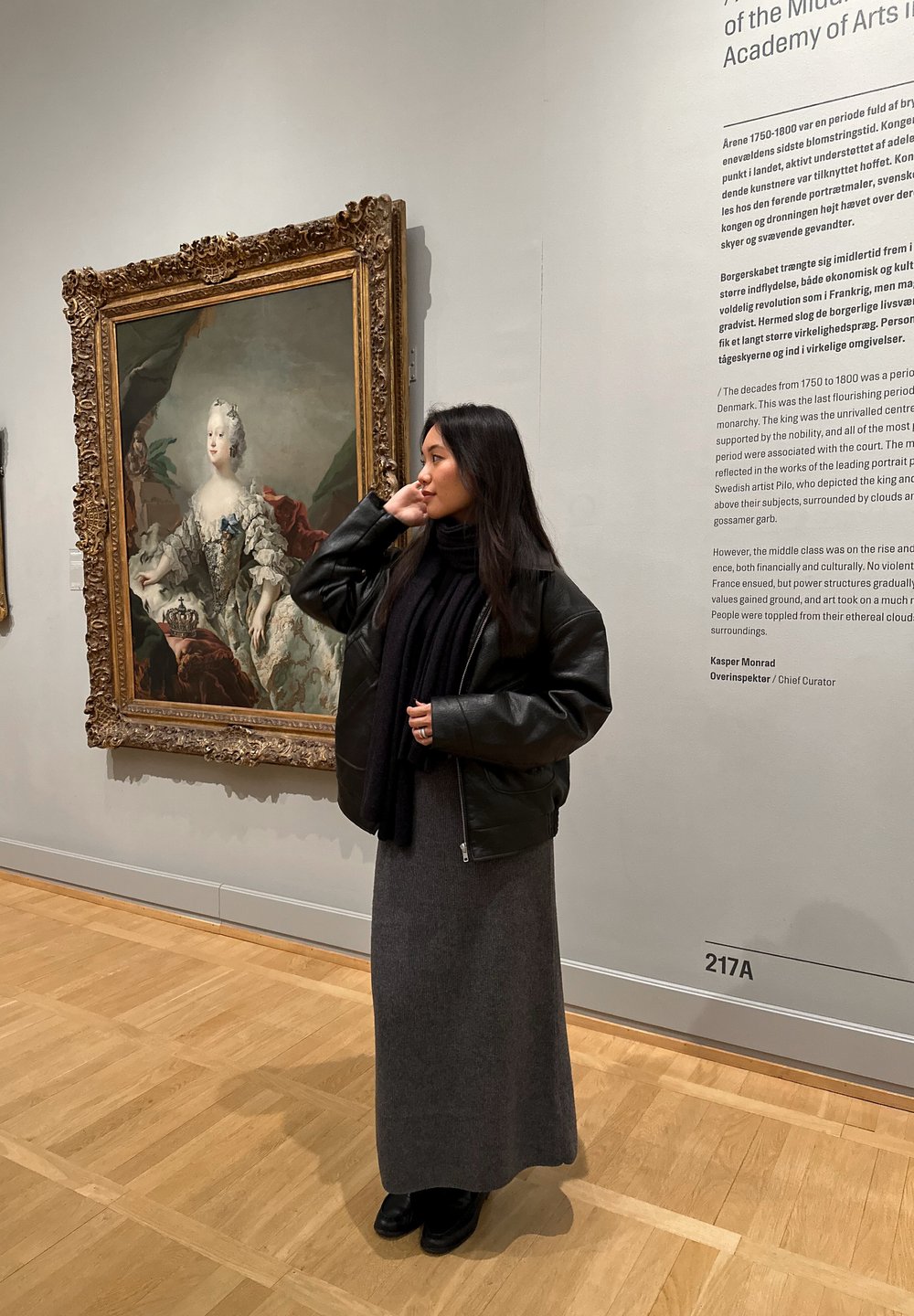 Osoba u crnoj kožnoj jakni preko tamno sive dugačke haljine stoji unutra pored okvira slike na svijetlom zidu.