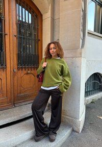 Olivengrønn oversized genser med vide ermer, kombinert med mørkebrune treningsbukser med gullstriper. Inkluderer brun veske og fottøy.