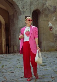 Rožinė švarkas, baltas marškinėliai su braškių piešiniu, raudoni plačiapakšiai kelnes, raudoni saulės akiniai ir grietinėlės spalvos rankinė. Vieta - akmenimis grįsta gatvė.