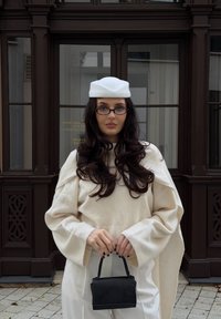Kremowy sweter z szerokimi rękawami, czarna torebka, biała beretka, długie ciemne włosy i okulary, w tle z oknem w panelach.