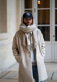 Cappotto di lana beige con due bottoni, indossato sopra una felpa grigia. Sciarpa grigia chiara con frange. Cappellino da baseball scuro con logo.
