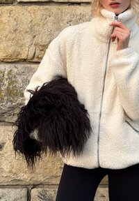 Giacca a zip in pile bianco con collo alto, caratterizzata da una finitura strutturata. Il modello tiene un accessorio in pelliccia sintetica nera con fili lunghi.