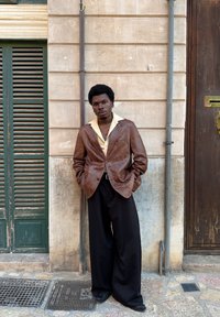 Blazer de couro castanho por cima de uma camisa amarelo-claro, combinado com calças pretas largas. De pé contra uma parede bege com persianas verdes.