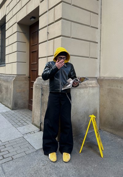 Chaqueta de cuero negra, sudadera con capucha amarilla, pantalones anchos negros, zapatillas amarillas y una revista en mano, cerca de una señal de advertencia amarilla.