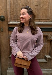 Femme souriante regardant vers la gauche, portant une chemise à manches longues rayée, un pantalon bordeaux, des bijoux en or, tenant un sac en daim marron devant une porte en bois.