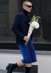 Person klädd i en skräddarsydd marinblå kavaj över en polotröja, blå ullshorts, som håller en bukett inslagen i papper med handskrivna anteckningar.