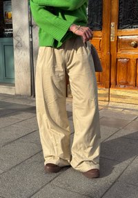 Άτομο που φοράει φαρδιά μπεζ παντελόνια, καφέ παπούτσια και φωτεινό πράσινο πουλόβερ, στέκεται στο πεζοδρόμιο κοντά σε μια ξύλινη πόρτα.