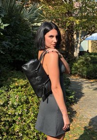 Schwarzer Leder-Rucksack mit verstellbaren Riemen, rechteckiger Form und strukturiertem Finish. Getragen von einer Person in einem gestreiften Kleid.