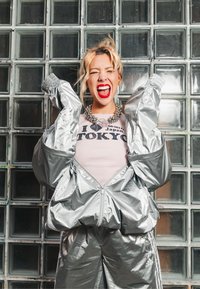 Survêtement argenté et réfléchissant avec une veste à fermeture éclair et un pantalon assorti, porté par-dessus un t-shirt rose pâle comportant un texte noir, devant un mur en briques de verre.