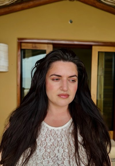 Primer plano de una mujer con cabello largo y oscuro que lleva una blusa de encaje blanca. El fondo muestra puertas de madera y un color de pared neutro.