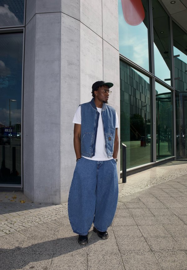 Mann mit schwarzer Kappe, Brille, weissem T-Shirt, blauer Jeansweste und weiten, baggy blauen Jeans steht vor einem modernen Glasgebäude.