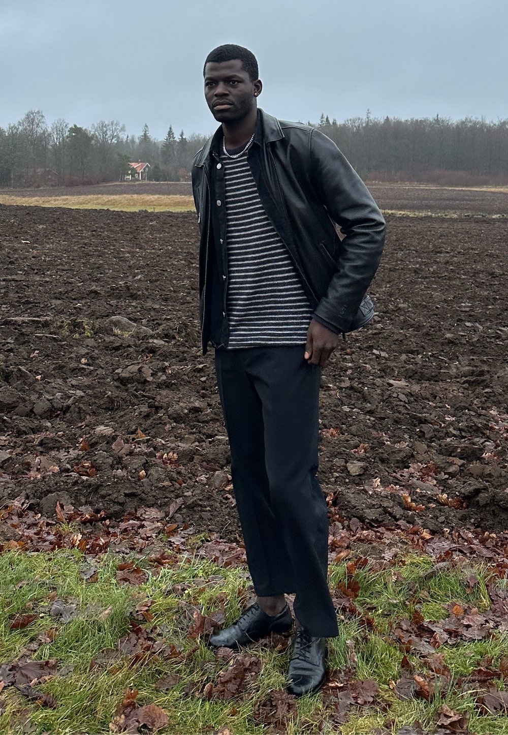 Homme portant une veste en cuir noire, une chemise rayée, un pantalon noir et des chaussures, debout sur l'herbe à côté d'un champ labouré avec des arbres en arrière-plan.