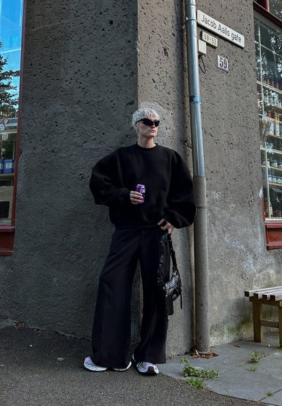 Sudadera negra de gran tamaño y pantalones negros de pierna ancha, combinados con zapatillas de deporte coloridas. Los accesorios incluyen un pequeño bolso negro y gafas de sol.