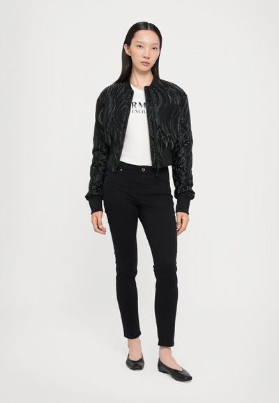 Chaqueta bomber negra con textura acolchada, cierre de cremallera y puños de canalé, llevada sobre una camiseta blanca y jeans ajustados negros. Bailarinas negras completan el look.