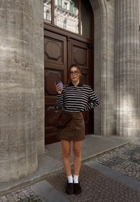 Ribade pikale mansetiga polo särk, pruun nahast seelik, pruun käekott, pruunad kingad, valged pahkluu sokid, hoides lillat tassi, puidust uste ees.