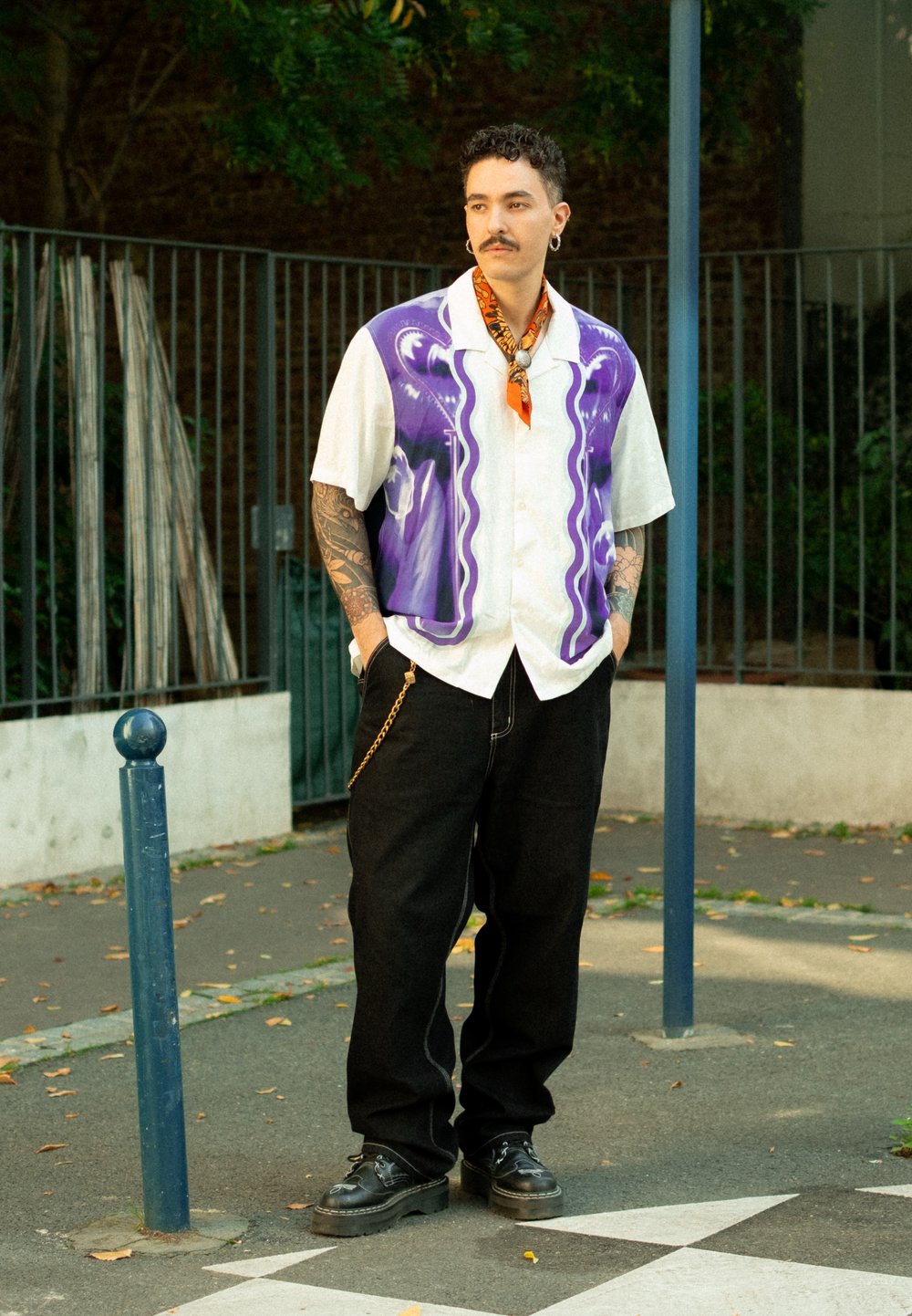 Man with curly hair and tattoos wears a purple patterned short-sleeve shirt, an orange scarf, black trousers, and platform shoes, standing outdoors.