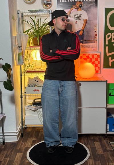 Camiseta negra de manga larga con rayas rojas, vaqueros baggy de color azul claro, zapatos redondos negros. Estanterías de fondo con plantas y zapatillas.