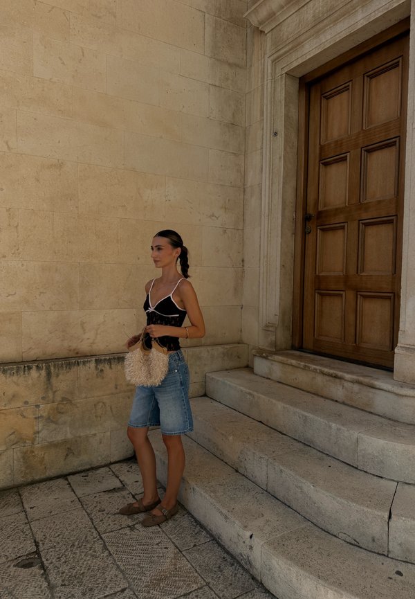 Femme en short en jean et haut noir tenant un sac à main beige debout près de marches en pierre et d'une grande porte en bois sur un mur en pierre beige.