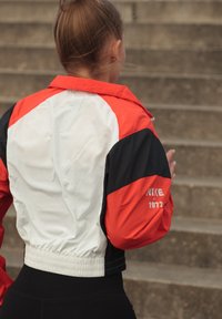 Ženska v rdeči, beli in črni jakni Nike z oznako "1972" na rokavu, ki hodi po zunanjih betonskih stopnicah, z lasmi vezanimi v punčo.
