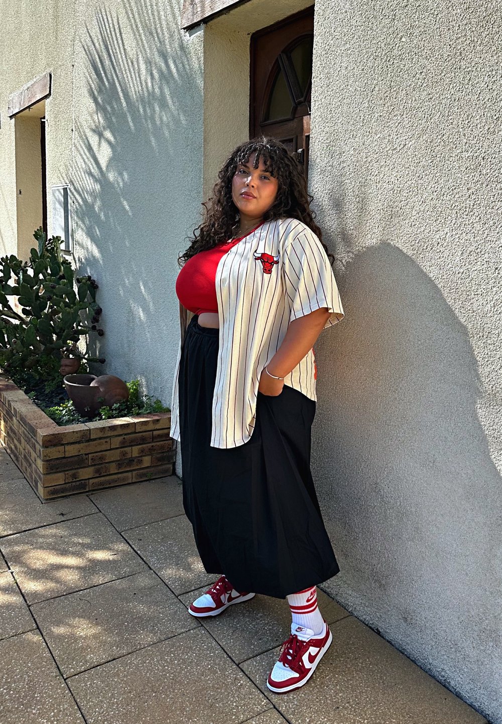 Camicia a maniche corte a righe con logo rosso, top rosso, gonna nera e sneaker rosso/bianco. Visibili vegetazione circostante e muro testurizzato.