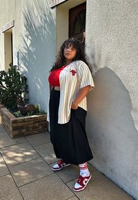Camicia a maniche corte a righe con logo rosso, top rosso, gonna nera e sneaker rosso/bianco. Visibili vegetazione circostante e muro testurizzato.
