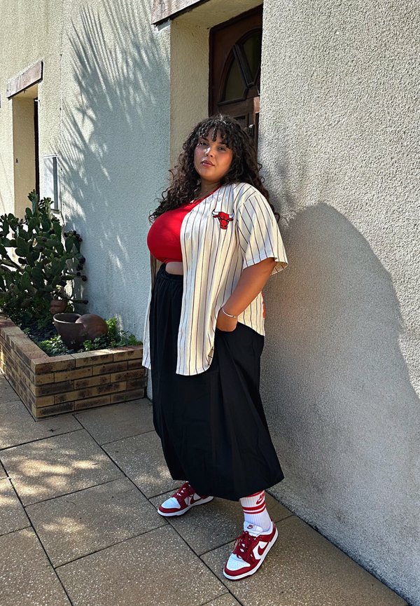 Camicia a maniche corte a righe con logo rosso, top rosso, gonna nera e sneaker rosso/bianco. Visibili vegetazione circostante e muro testurizzato.