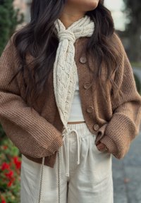 Hnědý pletený kardigan s velkými knoflíky, v kombinaci s texturovaným světle krémovým šátkem. Modelka má na sobě volné krémové kalhoty a přiléhavý top.