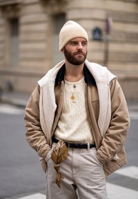 Beige Bomberjacke über weißem Strickpulli, beigen Hosen mit braunem Bandana, cremefarbenem Strickmütze, goldener Halskette und schwarzem Gürtel.