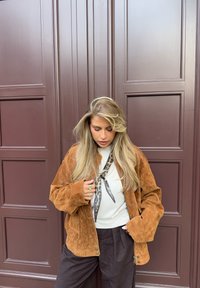 Brun semsket jakke med en avslappet passform, brukt over en hvit topp, kombinert med svarte bukser; inkluderer et mønstret skjerf og smykkedetaljer.