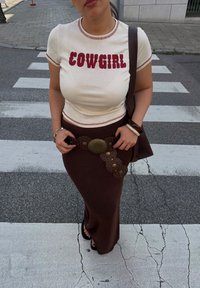 Cremefarvet t-shirt med "COWGIRL" i røde bogstaver, parret med en tekstureret brun midiskirt. Tilbehøret inkluderer et cirkulært mønstret bælte og en brun taske.