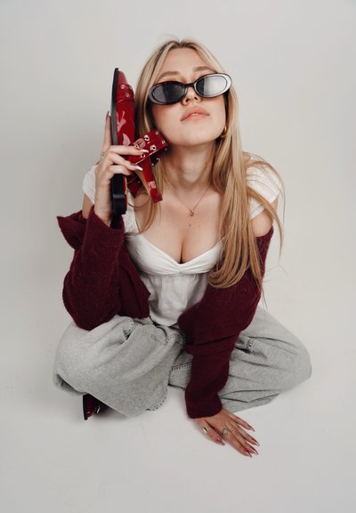 Zapatos de tacón alto rojos de charol sostenidos con una mano, combinados con jeans holgados grises y una blusa blanca, sobre la cual lleva un cárdigan de punto burdeos.