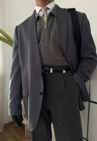 Harmaa räätälöity takki harmaan vetoketjullisen villapaidan päällä, yhdistettynä harmaisiin housuihin. Musta vyö ja hanskat tuovat kontrastia. Raidoitettu solmio näkyvissä.