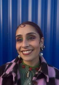 Personne souriante aux cheveux lissés en arrière, maquillage des yeux vert et violet, boucles d'oreilles en forme de cœur, colliers superposés et manteau à carreaux violet et noir sur fond bleu.