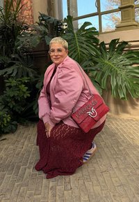 Giacca puffa rosa, gonna in pizzo bordeaux, borsa rossa con design "Coca-Cola" e sneakers a righe blu su uno sfondo di vegetazione interna.