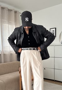 Čierna kožená bunda cez čierne polo tričko, béžové skladané nohavice a čierny klobúk s bielym logom, ktorý dopĺňa outfit čierny opasok.