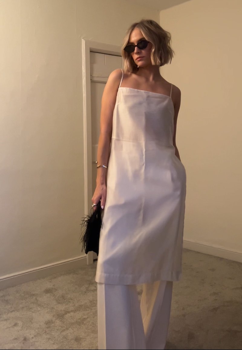 Femme aux cheveux blonds courts portant des lunettes de soleil noires, une robe blanche sans manches sur un pantalon blanc, tenant une pochette noire à plumes à l'intérieur.