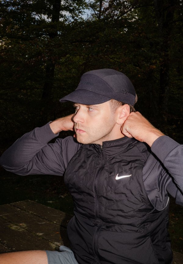 Hombre con chaleco negro de Nike y gorra oscura ajusta el cuello mientras está sentado en un banco al aire libre con árboles en el fondo al anochecer.