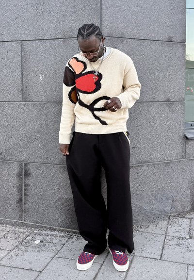 Hombre con suéter crema con diseño abstracto rojo y marrón, pantalones anchos negros, zapatillas rojas estampadas, de pie contra una pared de piedra gris.