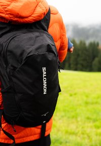 Person mit orangefarbener Jacke und schwarzem Salomon-Rucksack, der im Freien auf nassem Gras in der Nähe von Bäumen an einem bewölkten Tag steht.
