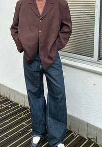 Blazer oversized castanho escuro com dois botões, combinado com jeans largos azuis escuros e sapatos pretos, com uma sola branca simples.
