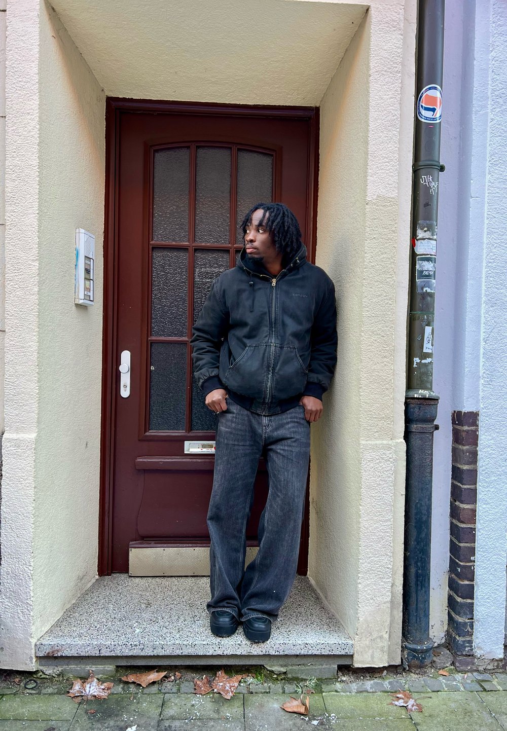 Homme en veste noire et jean appuyé contre un cadre de porte avec une porte marron, regardant sur le côté dans une rue parsemée de feuilles mortes.