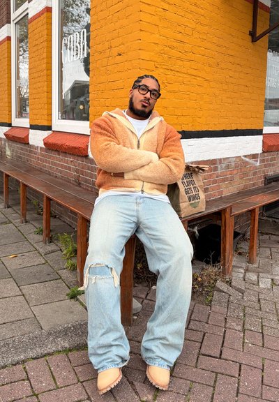 Hombre con gafas y cabello trenzado sentado en un banco frente a un edificio de ladrillos amarillos y blancos, vestido con una chaqueta marrón con cremallera y jeans rotos.